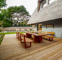 A modern outdoor wooden dining set with a rectangular table and matching benches on a large wooden deck, next to a house with a gray shingled roof, with a pool and trees in the background.